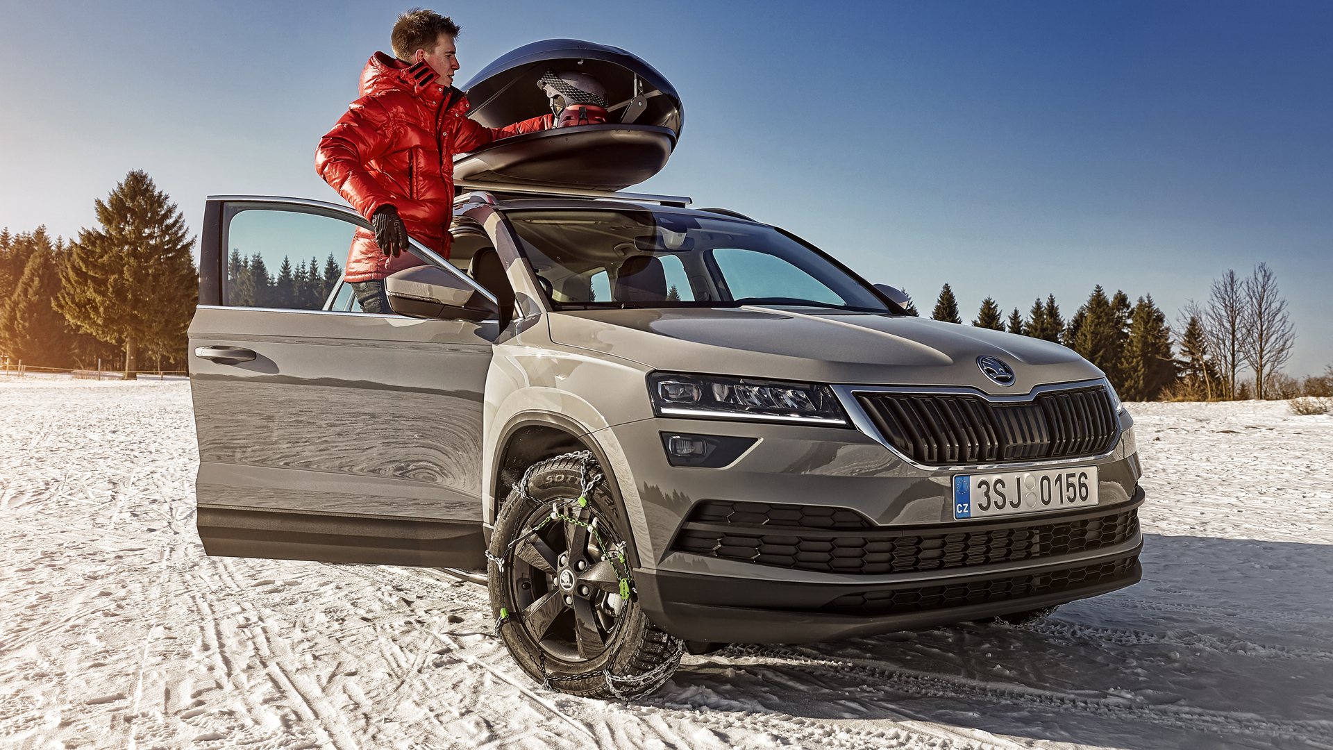 Homme chargeant un coffre de toit ouvert sur un Škoda Karoq gris stationné sur la neige