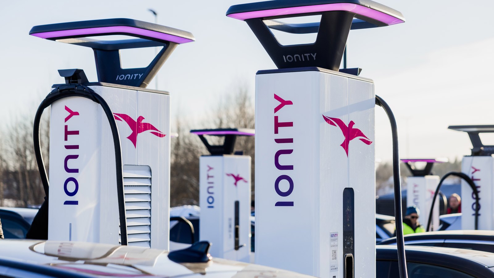 Bornes de recharge ultra-rapide IONITY blanches et roses alignées sur une station, voitures branchées en arrière-plan