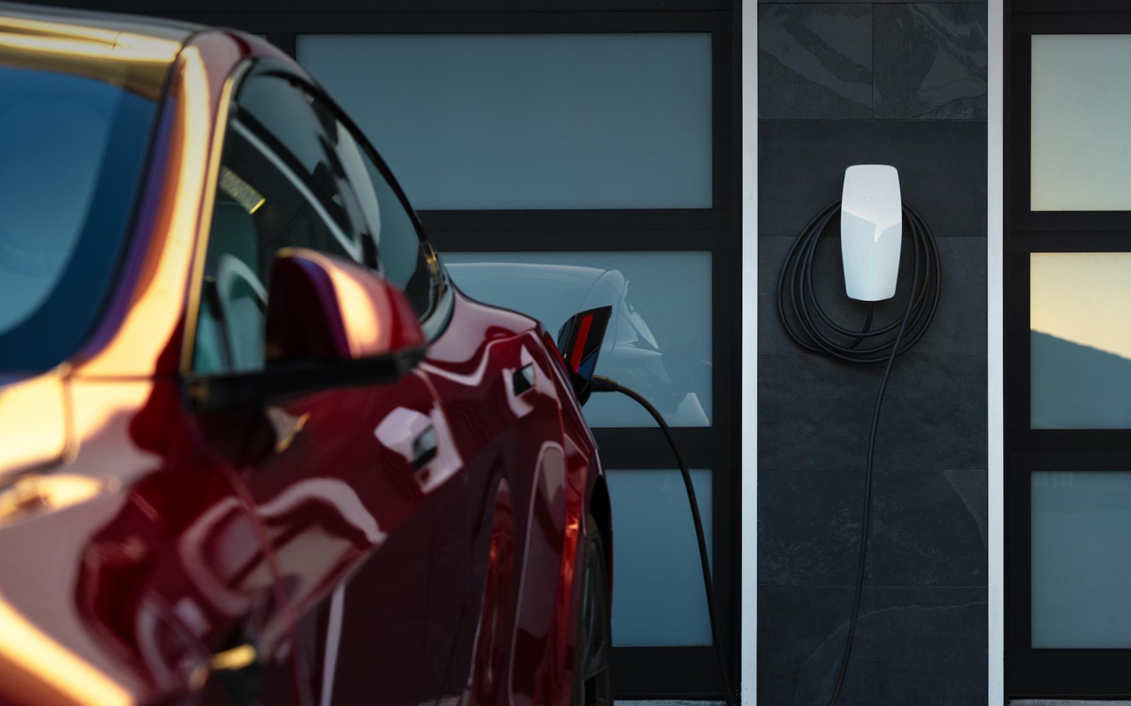 Tesla rouge branchée à un chargeur mural Tesla devant une maison, câble de recharge connecté