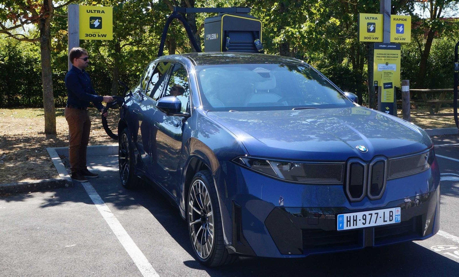 Une BMW iX3 bleue branchée à une borne ultra-rapide d'une station Le Plein, câble de recharge en main