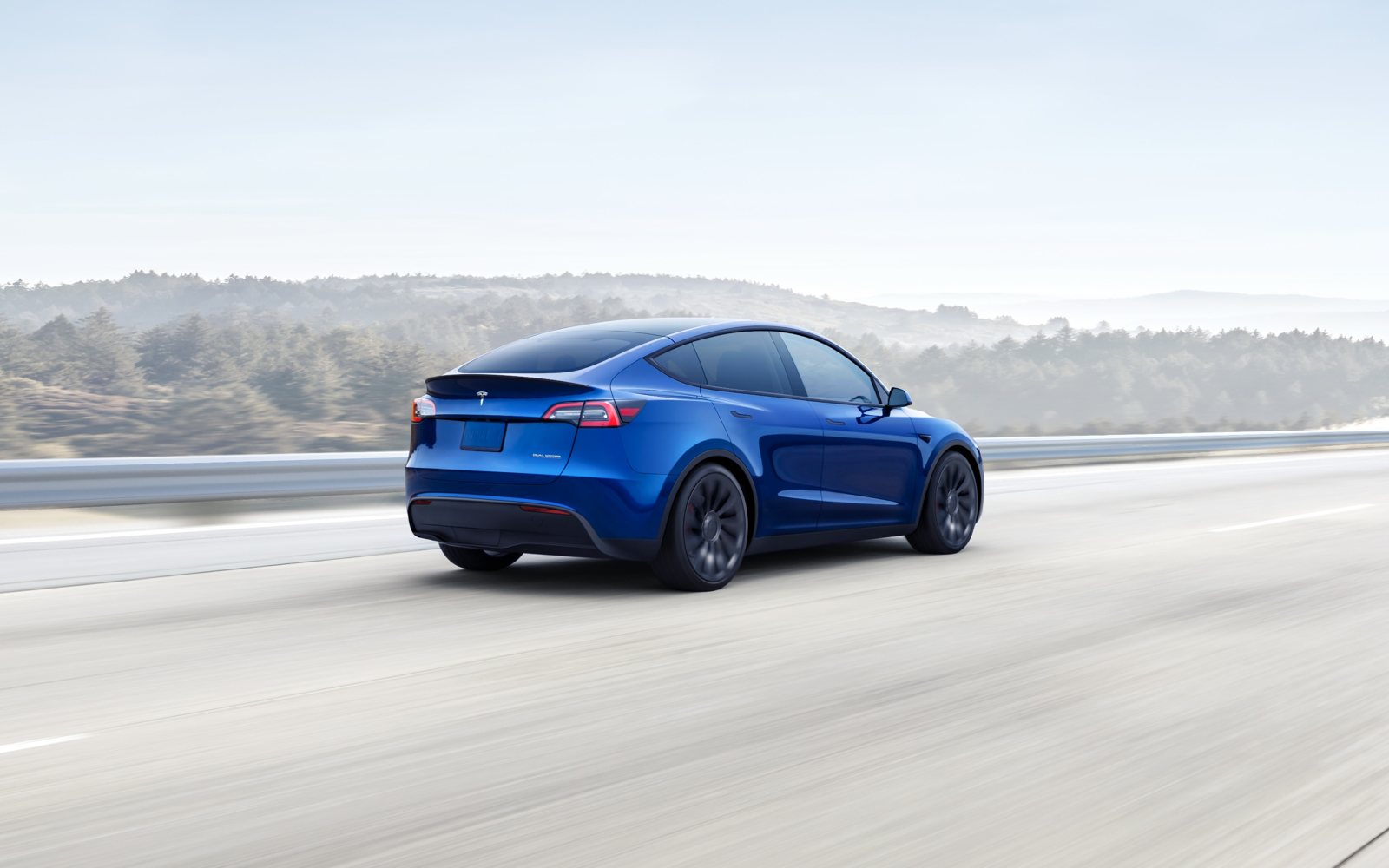 Tesla Model Y bleue vue de trois quarts arrière roulant sur une autoroute bordée de collines boisées